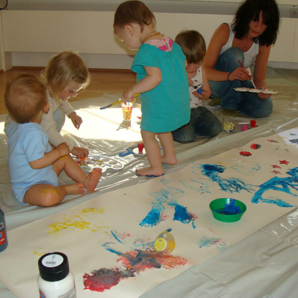 Mehrere kleine Kinder und ein Erwachsener sitzen auf dem Boden um ein großes Blatt Papier herum und malen mit den Fingern in verschiedenen Farben. Künstlerbedarf und Farbflaschen liegen verstreut in der Nähe.