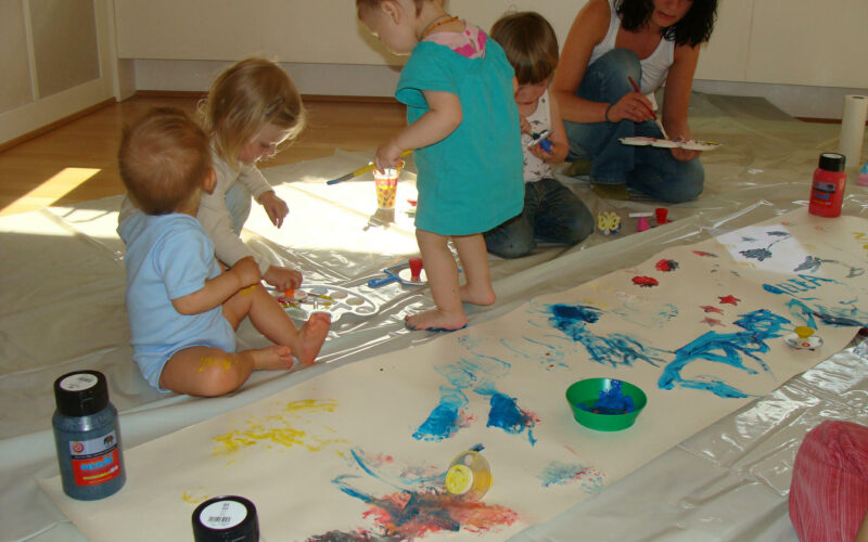 Mehrere kleine Kinder und ein Erwachsener sitzen auf dem Boden um ein großes Blatt Papier herum und malen mit den Fingern in verschiedenen Farben. Künstlerbedarf und Farbflaschen liegen verstreut in der Nähe.
