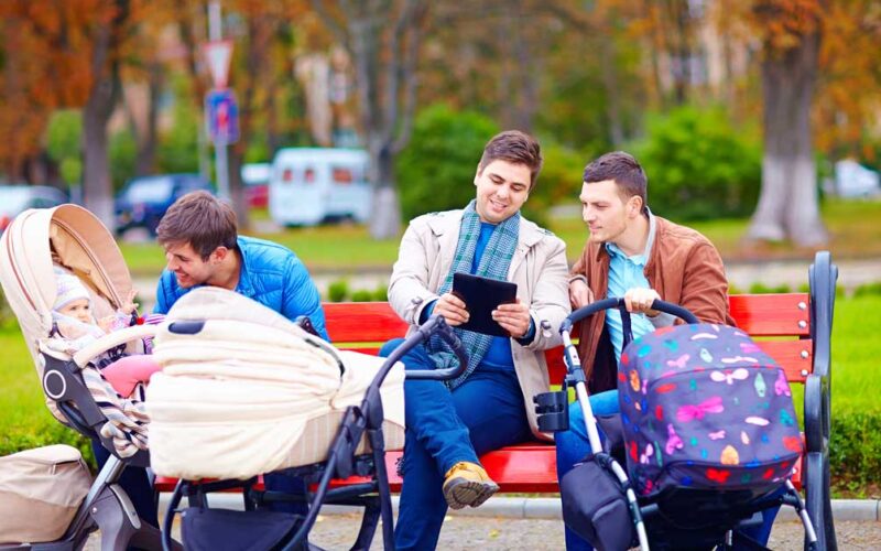 Drei Männer sitzen auf einer Parkbank, zwei mit Kinderwagen, einer mit einem Tablet. Ein Mann schaut auf ein Baby, während die anderen sich unterhalten. Im Hintergrund sind Herbstbäume zu sehen.