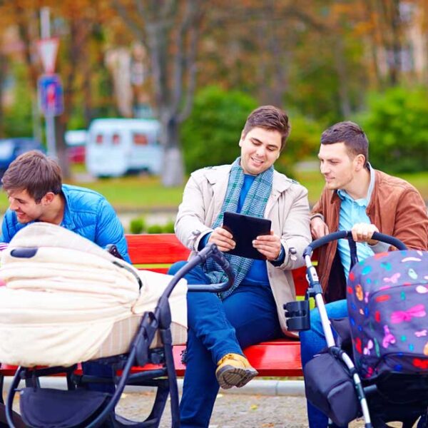 Drei Männer sitzen mit Kinderwagen auf einer Parkbank. Einer schaut nach einem Baby, ein anderer hält ein Tablet in der Hand, und der dritte blickt zur Gruppe. Im Hintergrund sind Bäume und geparkte Autos zu sehen.