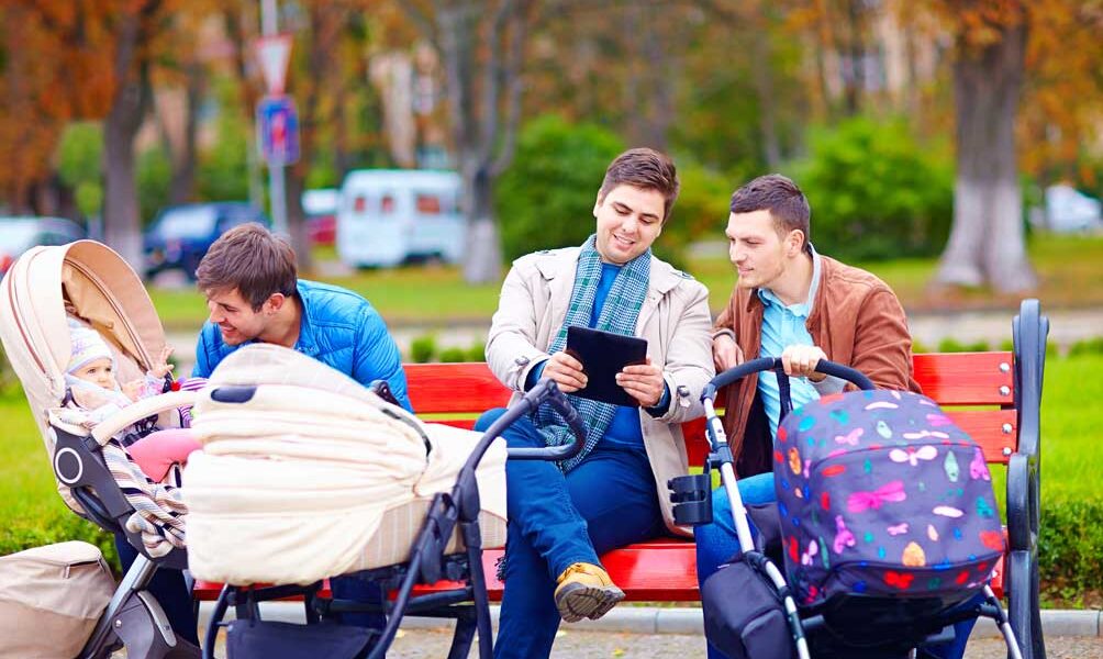 Drei Männer sitzen auf einer Parkbank mit zwei Kinderwagen; ein Mann benutzt ein Tablet, einer sieht nach einem Baby in einem Kinderwagen, und der dritte sitzt neben einem anderen Kinderwagen. Im Hintergrund sind Bäume und Autos zu sehen.