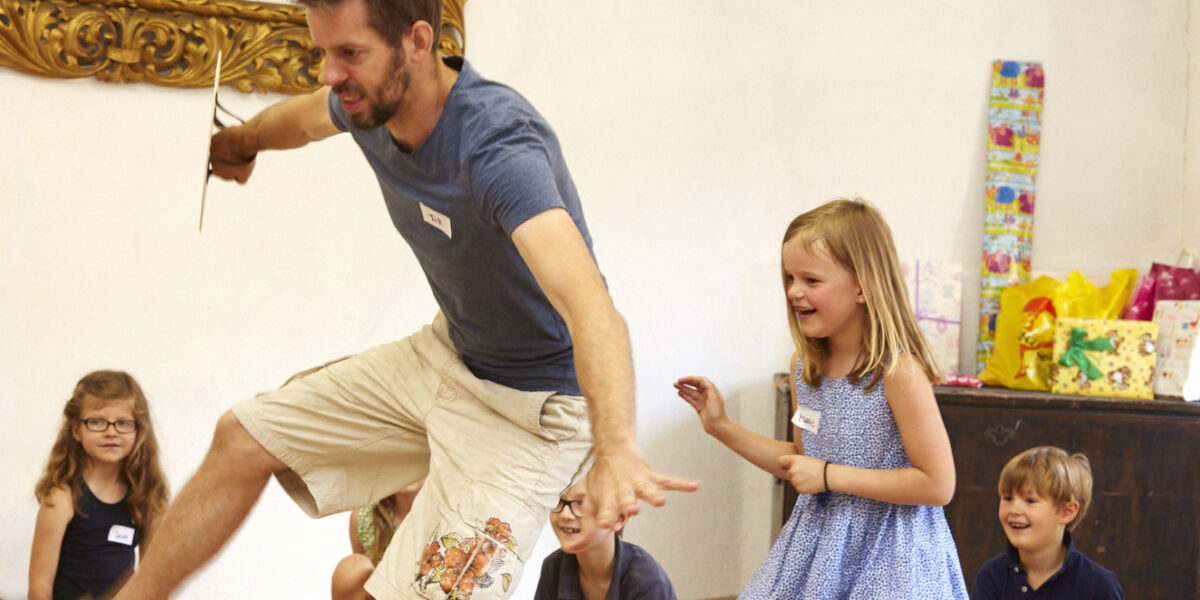 Ein erwachsener Mann springt spielerisch vor sitzenden und lachenden Kindern in einem Raum mit einer dekorierten Wand und Geschenken auf einem Schrank im Hintergrund.