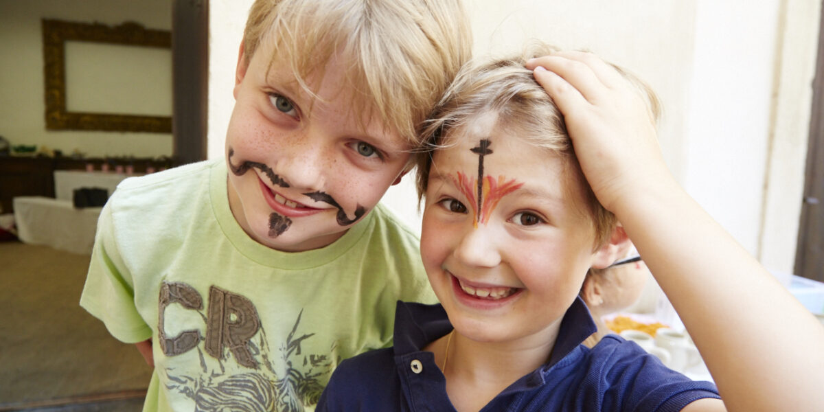 Zwei kleine Jungen lächeln in die Kamera. Der eine hat einen aufgemalten Schnurrbart und einen Bart, der andere ist über den Augenbrauen geschminkt und berührt seinen Kopf.