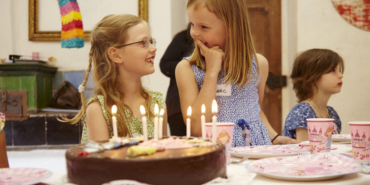 Drei Kinder sitzen an einem Tisch mit Schokoladengeburtstagstorte, Kerzen und rosa Partyzubehör. Zwei Mädchen lächeln und unterhalten sich, während ein Junge daneben sitzt.