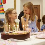 Drei Kinder sitzen an einem Tisch mit Schokoladengeburtstagstorte, Kerzen und rosa Partyzubehör. Zwei Mädchen lächeln und unterhalten sich, während ein Junge daneben sitzt.