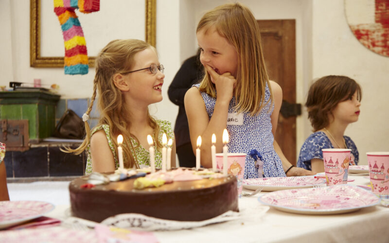 Drei Kinder sitzen an einem Tisch mit Schokoladengeburtstagstorte, Kerzen und rosa Partyzubehör. Zwei Mädchen lächeln und unterhalten sich, während ein Junge daneben sitzt.