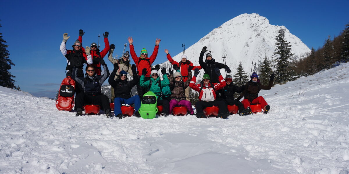 Eine Gruppe von Menschen in Winterkleidung sitzt mit erhobenen Armen auf Schlitten im Schnee, im Hintergrund sind ein schneebedeckter Berg und Bäume zu sehen.