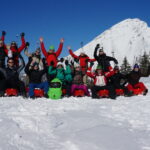 Eine Gruppe von Menschen in Winterkleidung sitzt mit erhobenen Armen auf Schlitten im Schnee, im Hintergrund sind ein schneebedeckter Berg und Bäume zu sehen.