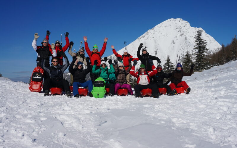 Eine Gruppe von Menschen in Winterkleidung sitzt mit erhobenen Armen auf Schlitten im Schnee, im Hintergrund sind ein schneebedeckter Berg und Bäume zu sehen.