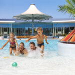 Vier Kinder spielen und planschen in einem Außenschwimmbad in der Nähe einer bunten Struktur und einer Palme, mit einem modernen Gebäude und Sonnenschirm im Hintergrund.