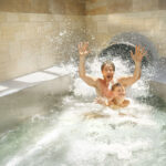 Ein Mann und ein Kind spritzen in ein Becken am Ende einer Indoor-Wasserrutsche, beide lächeln und heben ihre Hände, während das Wasser um sie herum spritzt.