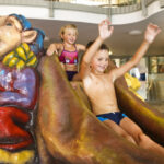 Zwei Kinder rutschen eine Wasserrutsche in Form einer skurrilen Figur in einem Hallenbad hinunter.