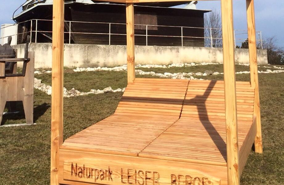 Eine große Lounge-Sitzgruppe aus Holz im Freien mit einer Pergola-Struktur, beschriftet mit „Naturpark Leiser Berge“, auf Gras gelegen, mit einem Holzturm im Hintergrund.