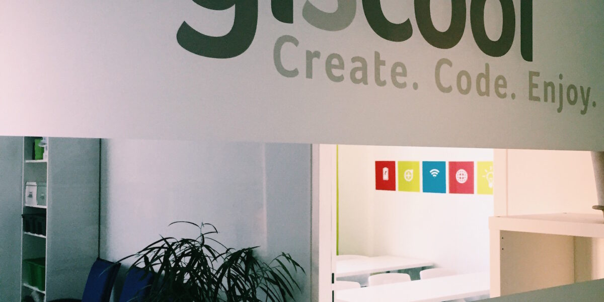 Glaswand mit dem Logo "logiscool" und dem Slogan "Create. Code. Enjoy." in einem Büroraum mit Stühlen, einer Pflanze und bunten Symbolen an der Rückwand.