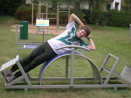 Eine Person liegt seitlich auf einem gebogenen Trainingsgerät im Freien auf einer Parkwiese, die Hände hinter dem Kopf und die Beine ausgestreckt.