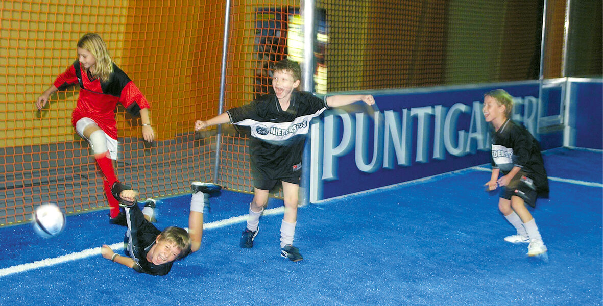 Vier Kinder spielen Hallenfußball; eines in Rot schießt den Ball in die Nähe des Tores, während drei in Schwarz reagieren, wobei eines zu Boden fällt.