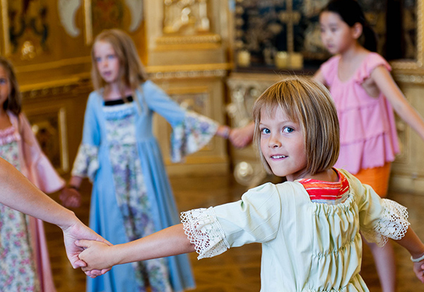Fünf kleine Kinder, einige davon in historischen Kostümen, bilden in einem reich verzierten Raum mit Golddekor einen Kreis an den Händen.