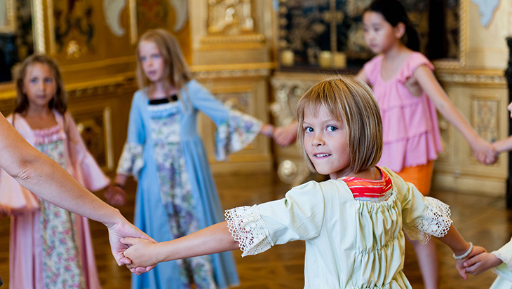 Fünf kleine Kinder, einige davon in historischen Kostümen, bilden in einem reich verzierten Raum mit Golddekor einen Kreis an den Händen.