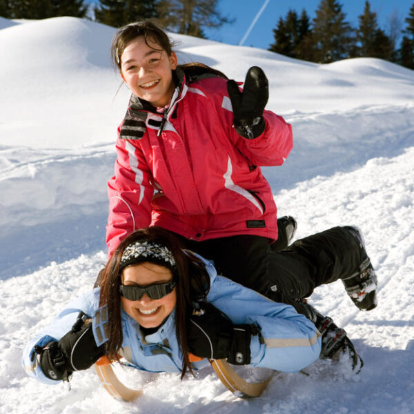 Zwei Mädchen fahren mit dem Schlitten einen verschneiten Hügel hinunter, wobei die eine auf dem Schlitten liegt und die andere auf ihrem Rücken fährt. Beide sind in Winterkleidung gekleidet und lächeln. Im Hintergrund sind Tannenbäume zu sehen.