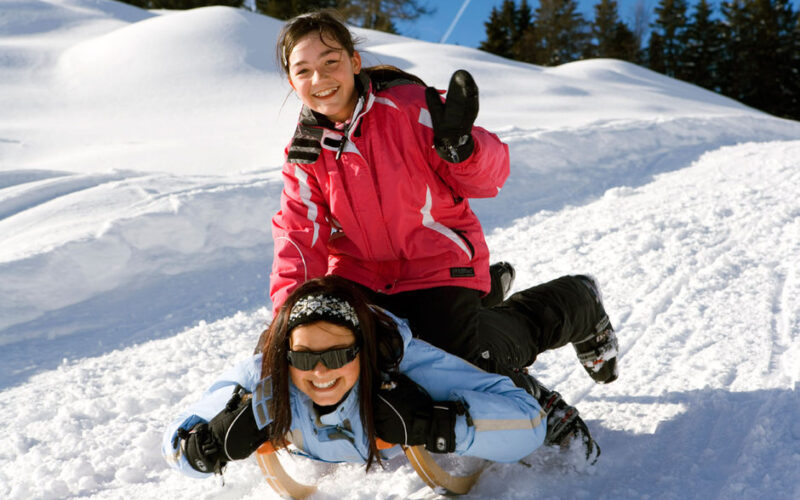 Zwei Mädchen fahren mit dem Schlitten einen verschneiten Hügel hinunter, wobei die eine auf dem Schlitten liegt und die andere auf ihrem Rücken fährt. Beide sind in Winterkleidung gekleidet und lächeln. Im Hintergrund sind Tannenbäume zu sehen.