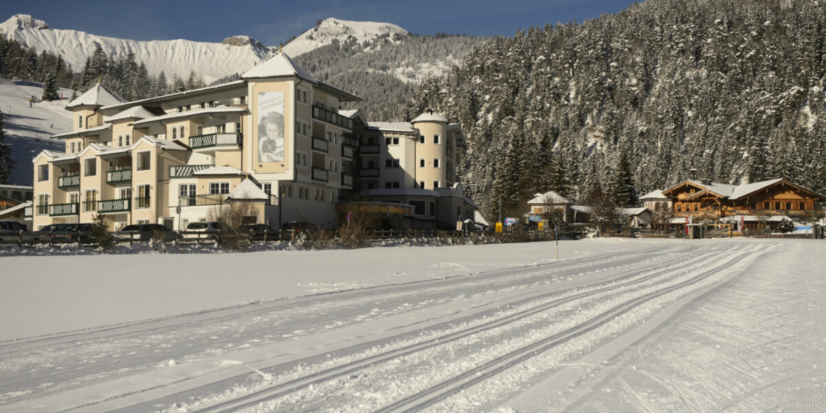 Ein großes Hotel steht neben hölzernen Gebäuden am Rande einer schneebedeckten Landschaft, mit Spuren im Schnee und bewaldeten Bergen im Hintergrund.