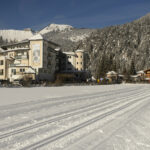 Ein großes Hotel steht neben hölzernen Gebäuden am Rande einer schneebedeckten Landschaft, mit Spuren im Schnee und bewaldeten Bergen im Hintergrund.