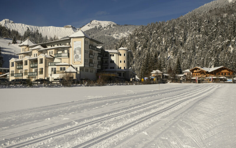 Ein großes Hotel steht neben hölzernen Gebäuden am Rande einer schneebedeckten Landschaft, mit Spuren im Schnee und bewaldeten Bergen im Hintergrund.