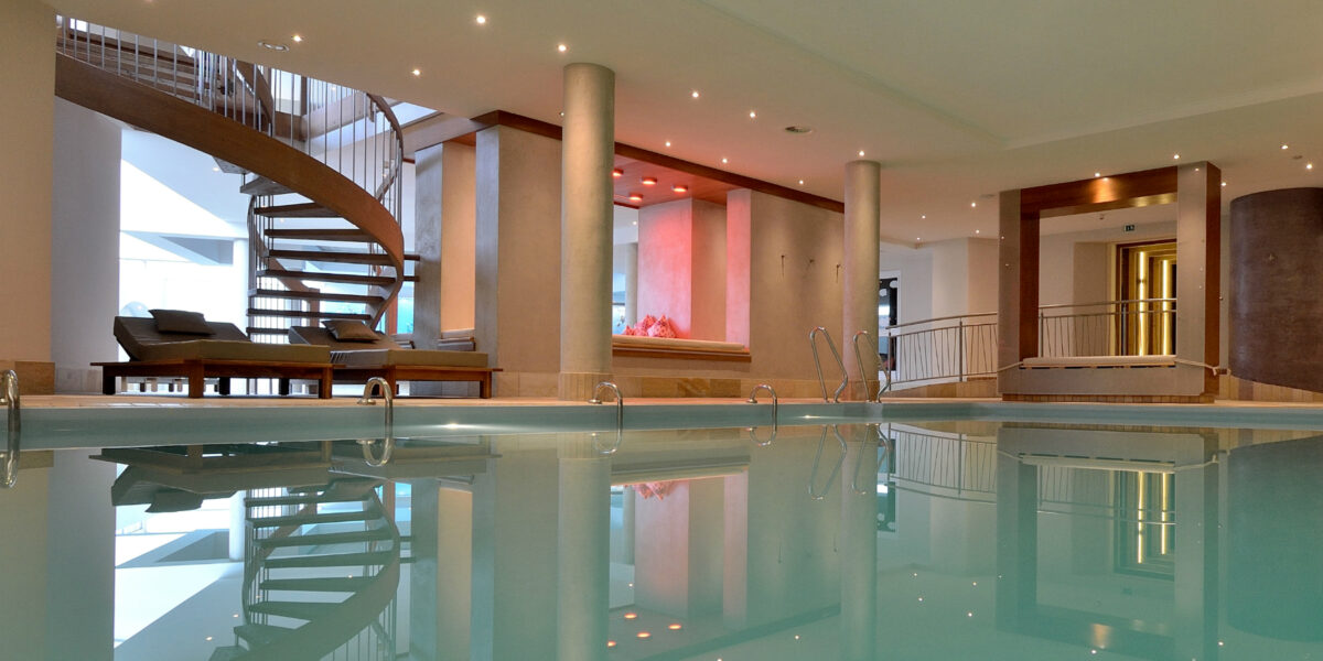 Hallenbadbereich mit Liegestühlen, Säulenstützen, einer Wendeltreppe und einer Deckenbeleuchtung, die sich im Wasser spiegelt.