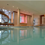 Hallenbadbereich mit Liegestühlen, Säulenstützen, einer Wendeltreppe und einer Deckenbeleuchtung, die sich im Wasser spiegelt.