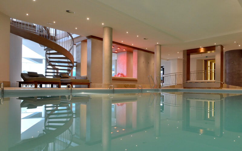 Hallenbadbereich mit Liegestühlen, Säulenstützen, einer Wendeltreppe und einer Deckenbeleuchtung, die sich im Wasser spiegelt.