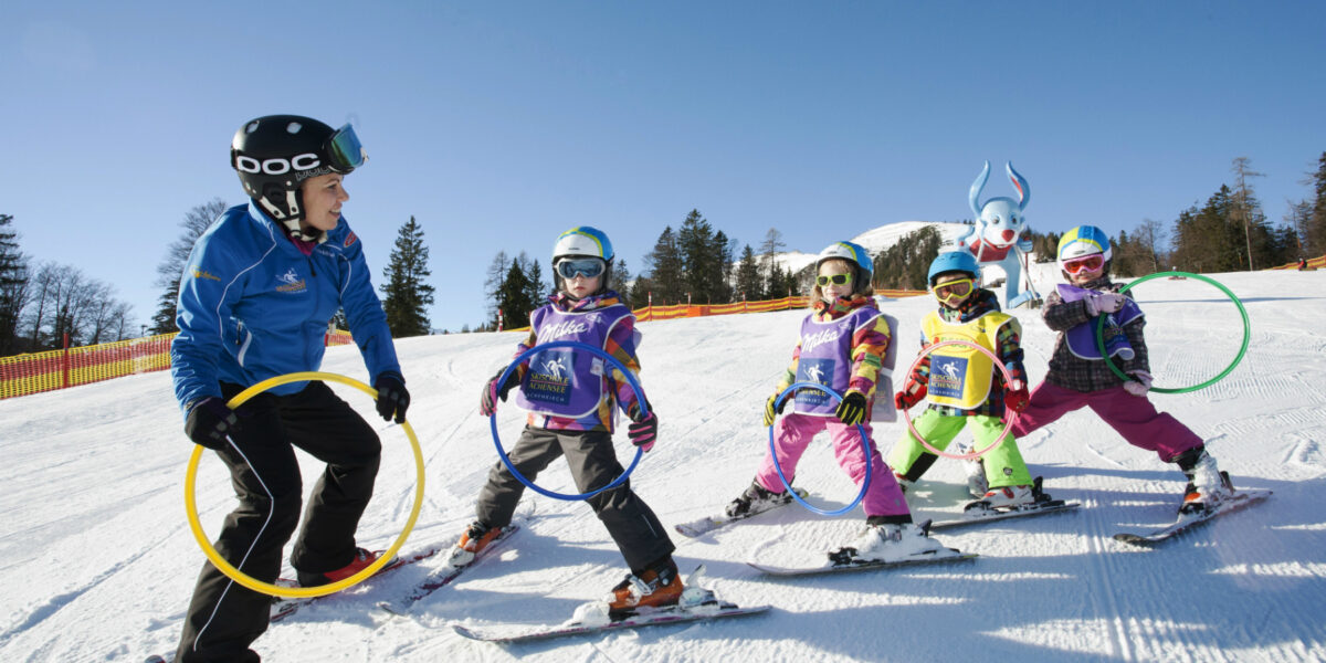 Ein Skilehrer hält Reifen und führt vier Kinder auf Skiern, während sie auf einer verschneiten Piste unter blauem Himmel üben.