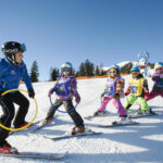 Ein Skilehrer hält Reifen und führt vier Kinder auf Skiern, während sie auf einer verschneiten Piste unter blauem Himmel üben.