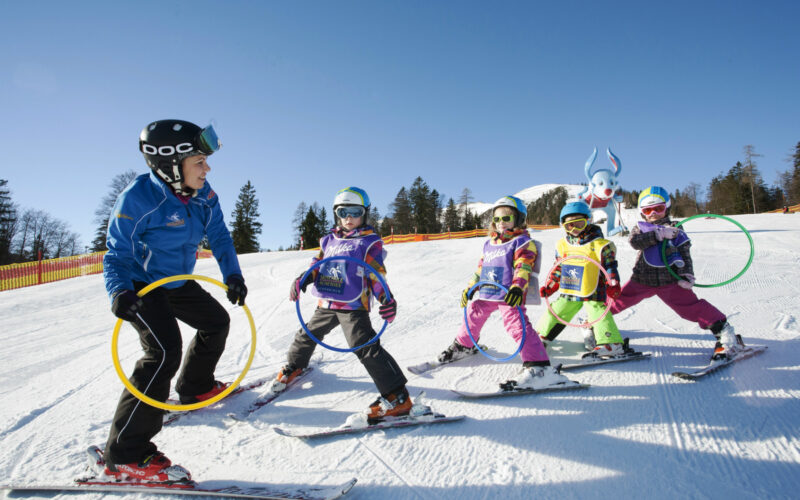 Ein Skilehrer hält Reifen und führt vier Kinder auf Skiern, während sie auf einer verschneiten Piste unter blauem Himmel üben.