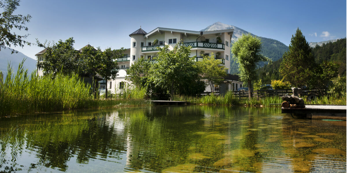Ein weißes Gebäude mit Balkonen steht neben einem Teich mit klarem Wasser und umliegendem Grün, vor einer Bergkulisse unter blauem Himmel.