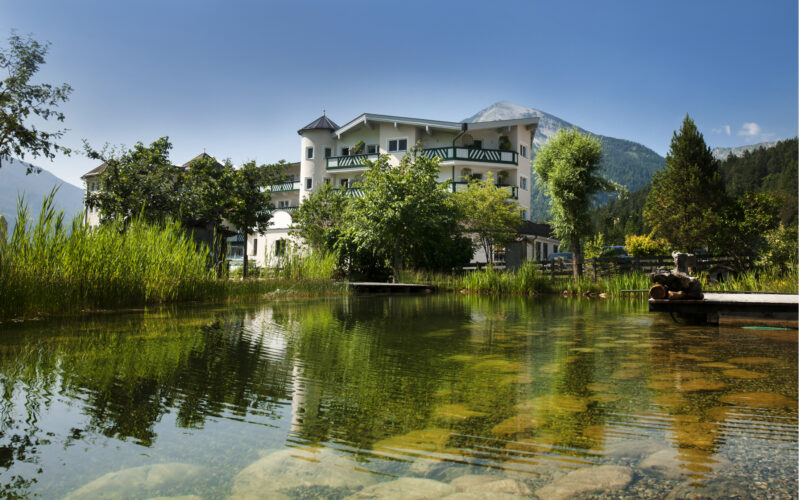 Ein weißes Gebäude mit Balkonen steht neben einem Teich mit klarem Wasser und umliegendem Grün, vor einer Bergkulisse unter blauem Himmel.