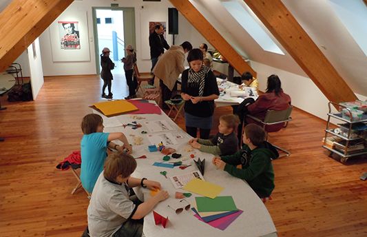 In einem dachbodenähnlichen Raum sitzen Kinder und Erwachsene an Tischen und beschäftigen sich mit buntem Papier und anderen Materialien.