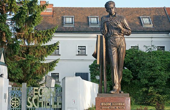 Vor einem weißen Gebäude mit Ziegeldach und Tor steht auf einem Sockel mit der Aufschrift „Egon Schiele“ eine bronzene Männerstatue.