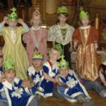 Eine Gruppe von Kindern in mittelalterlichen Kostümen und mit Papierkronen posiert gemeinsam im Innenbereich, manche sitzend, manche stehend.