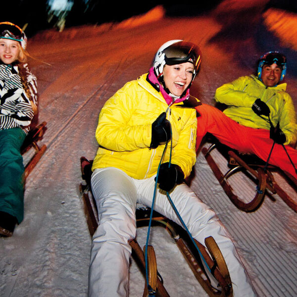 Drei Personen in Winterkleidung fahren nachts auf Holzschlitten einen verschneiten Hang hinunter. In der unteren rechten Ecke steht auf einem Banner „7 km Rodelspaß! Rodelspaß!“.