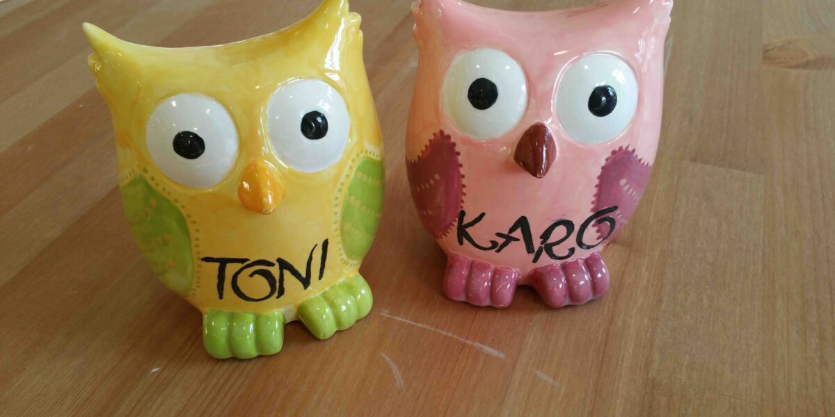 Zwei Eulenfiguren aus Keramik auf einer hölzernen Unterlage; die gelbe ist mit "TONI" und die rosa mit "KARO" beschriftet.