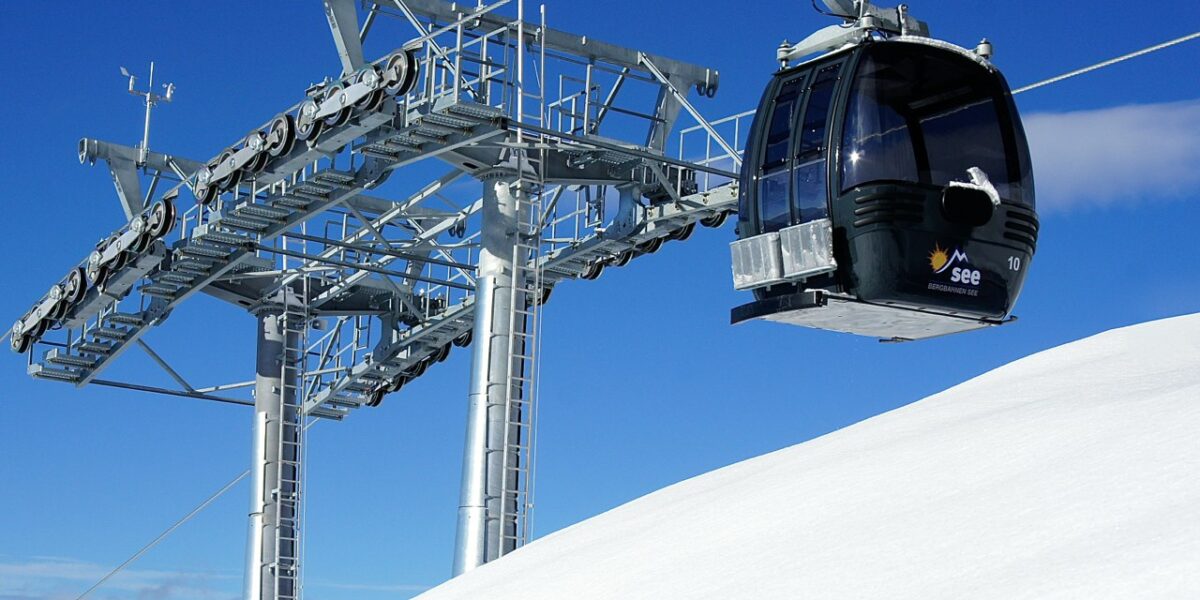 Eine Gondelbahn befördert die Fahrgäste über einen verschneiten Berghang, wobei die Stütztürme und ein Windsack unter einem klaren blauen Himmel zu sehen sind.