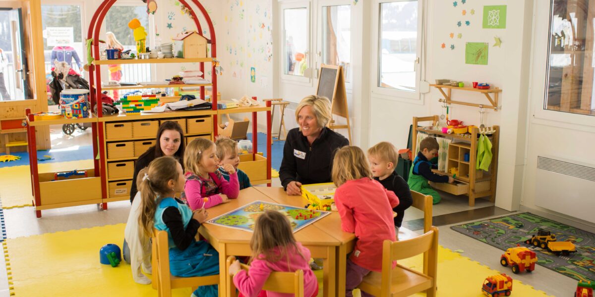 Eine Gruppe kleiner Kinder sitzt mit zwei Erwachsenen in einem hell erleuchteten Klassenzimmer um einen Tisch und spielt mit Lernspielzeug und Spielen auf einer gelben Matte.