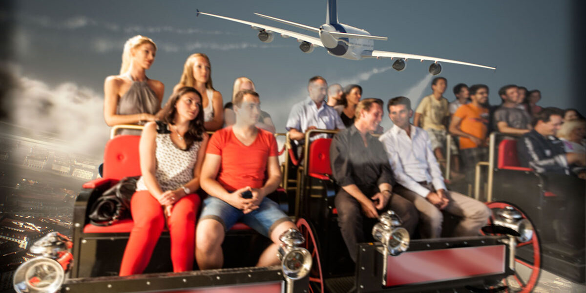 Eine Gruppe von Personen sitzt in einem Fahrsimulator und scheint eine Szene zu erleben, in der ein großes Flugzeug über dem Bildschirm im Hintergrund fliegt.