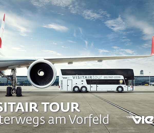 Ein geparktes Flugzeug auf der Rollbahn mit einem VISITAIR TOUR Bus daneben am Flughafen Wien, mit Terminalgebäuden im Hintergrund.