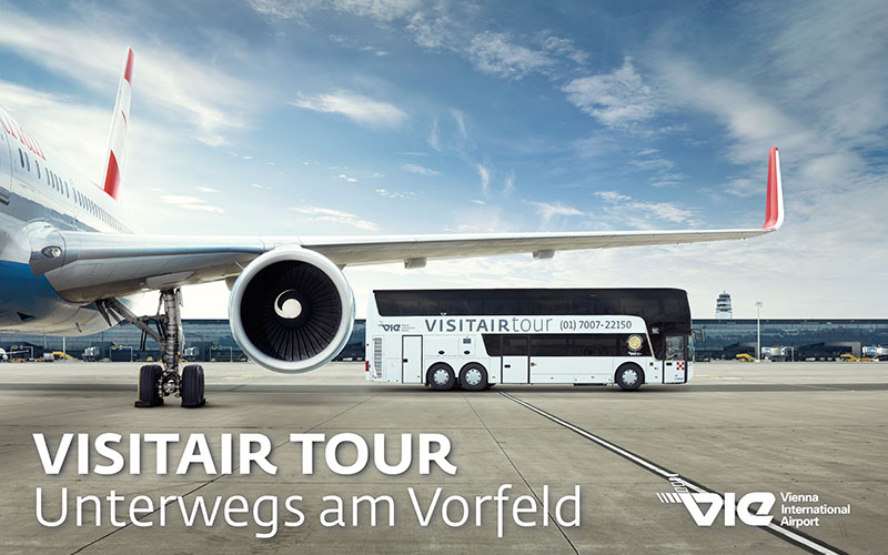 Ein geparktes Flugzeug auf der Rollbahn mit einem VISITAIR TOUR Bus daneben am Flughafen Wien, mit Terminalgebäuden im Hintergrund.