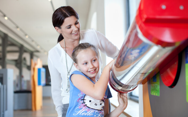 Eine Frau steht hinter einem lächelnden Mädchen, das in einem hell erleuchteten Wissenschaftsmuseum mit einem Exponat interagiert, das eine große rot-silberne Röhre zeigt.