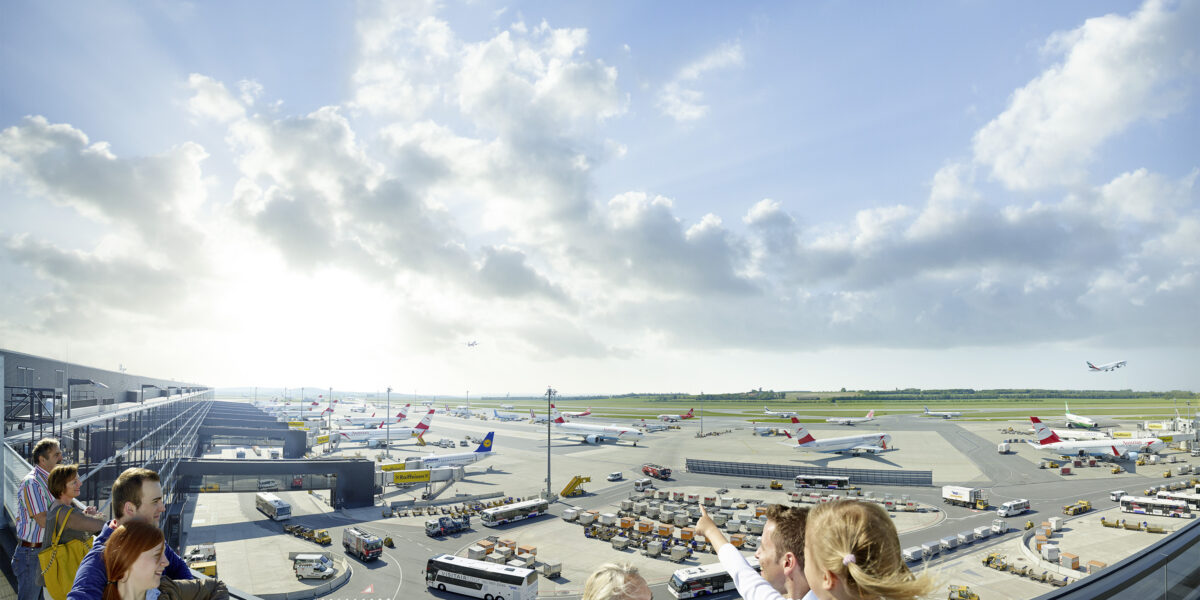 An einem sonnigen Tag mit teilweise bewölktem Himmel beobachten Menschen eine belebte Landebahn eines Flughafens mit mehreren geparkten Flugzeugen, Fahrzeugen und Geräten.