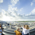 An einem sonnigen Tag mit teilweise bewölktem Himmel beobachten Menschen eine belebte Landebahn eines Flughafens mit mehreren geparkten Flugzeugen, Fahrzeugen und Geräten.