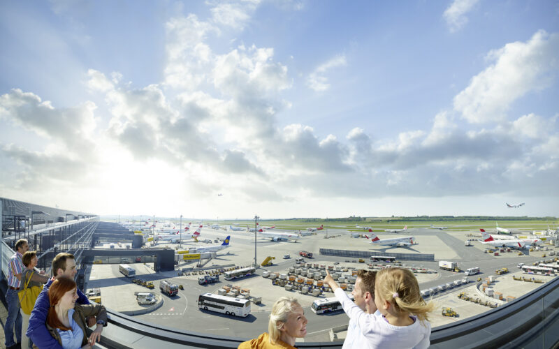 An einem sonnigen Tag mit teilweise bewölktem Himmel beobachten Menschen eine belebte Landebahn eines Flughafens mit mehreren geparkten Flugzeugen, Fahrzeugen und Geräten.