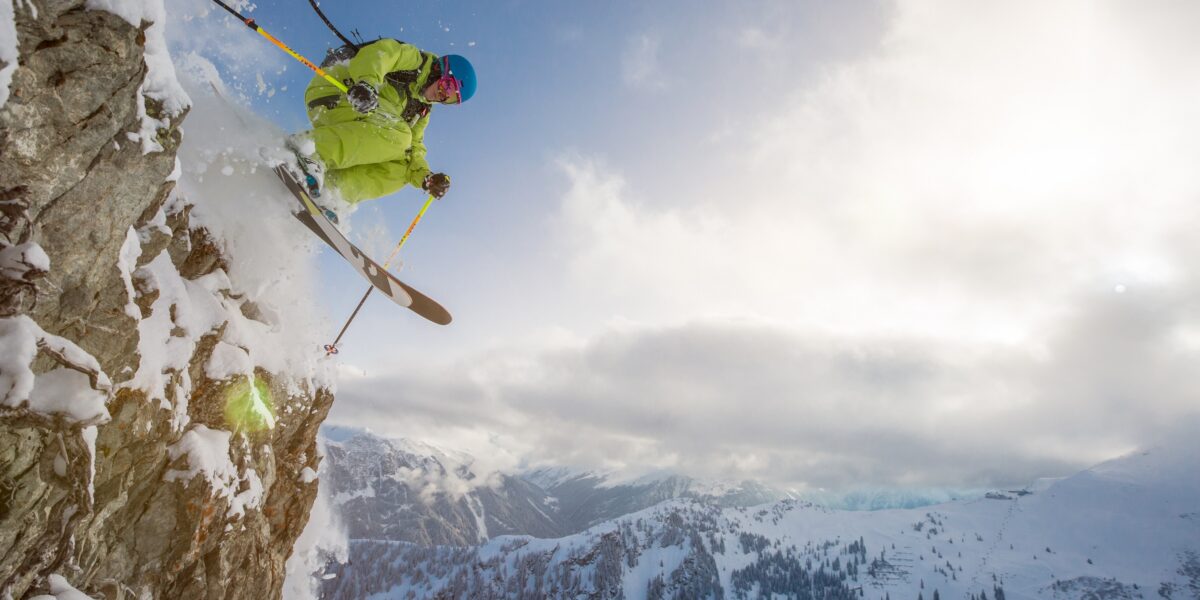 Ein Skifahrer in leuchtender Ausrüstung springt von einer schneebedeckten Klippe, im Hintergrund sind Berge und ein bewölkter Himmel zu sehen.